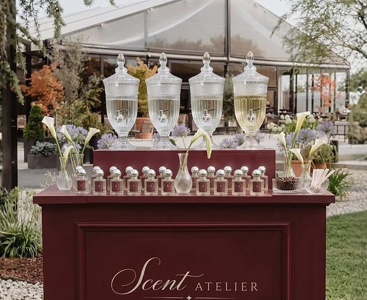 Parfemski bar Scent Atelier postavljen na travnjaku ispred šatora — četiri kristalna dozatora, niz bočica, kale i lavanda.